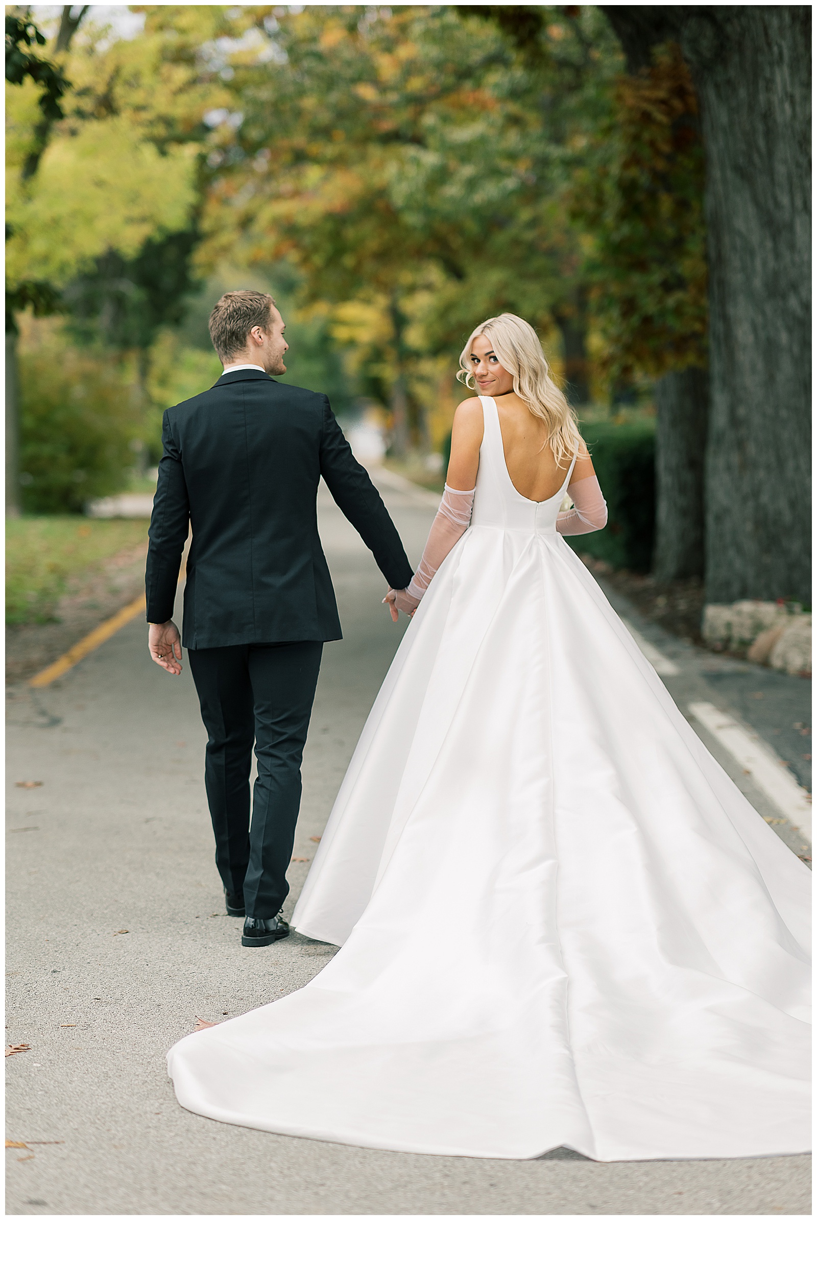 A bride and groom walk hand in hand with their backs turned down a narrow, empty street surrounded by a canopy of green leafy trees.  The bride looks back toward the camera with a soft smile.