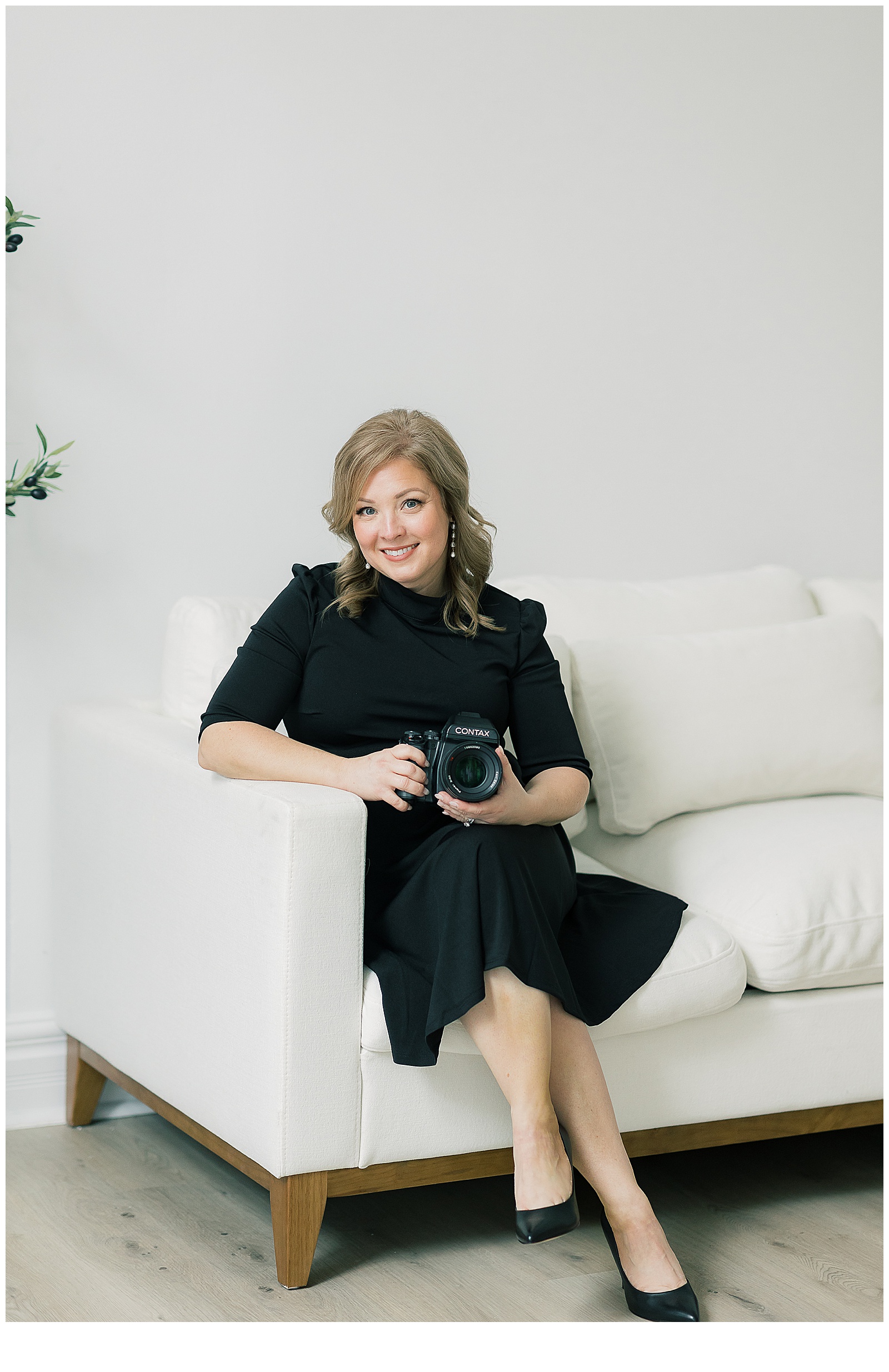 The photographer, Amy Simkus, is wearing a black dress and black high heels while seated on a modern white sofa holding a medium format film camera.